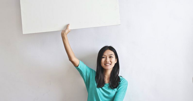 Real-Time Marketing - Cheerful Asian woman sitting cross legged on floor against white wall in empty apartment and showing white blank banner